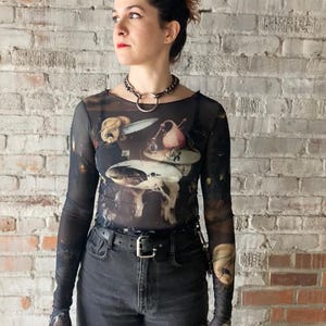 May include: A woman wearing a black mesh long-sleeved shirt with a painting of a still life on it. She is also wearing black jeans and a black choker necklace.
