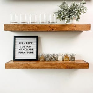 May include: Two wooden shelves with various items. The top shelf holds six clear glasses and a potted plant. The bottom shelf displays a letter board with text and small glass jars of spices. The letter board reads "HRNTREE CUSTOM HANDMADE FURNITURE."