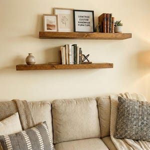 May include: Two rustic, wooden floating shelves mounted on a cream-coloured wall. The top shelf holds framed art, books, and a small plant. The bottom shelf displays books and a decorative vase. The text on the sign reads "IIRNTREE CUSTOM HANDMADE FURNITURE."