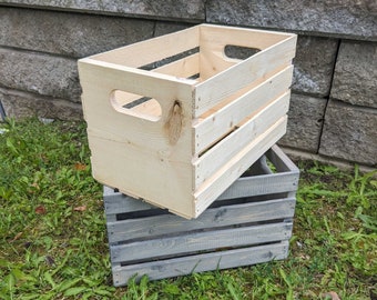 May include: Two wooden crates, one stacked on top of the other, sit on a bed of green grass. The top crate is unfinished, light-colored wood with hand-holds. The bottom crate is painted a muted gray.