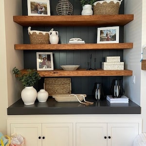 May include: A built-in shelving unit with dark wood shelves and a dark blue-green backing. The shelves display various decorative items, including framed photos, white vases, woven baskets, and decorative boxes. The unit is built over a white cabinet.