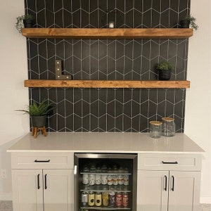 May include: A home bar with a black tiled feature wall and wooden shelves. The bar includes a mini-fridge filled with drinks, a white countertop, and grey cabinets. Decorative plants and glass jars enhance the design.