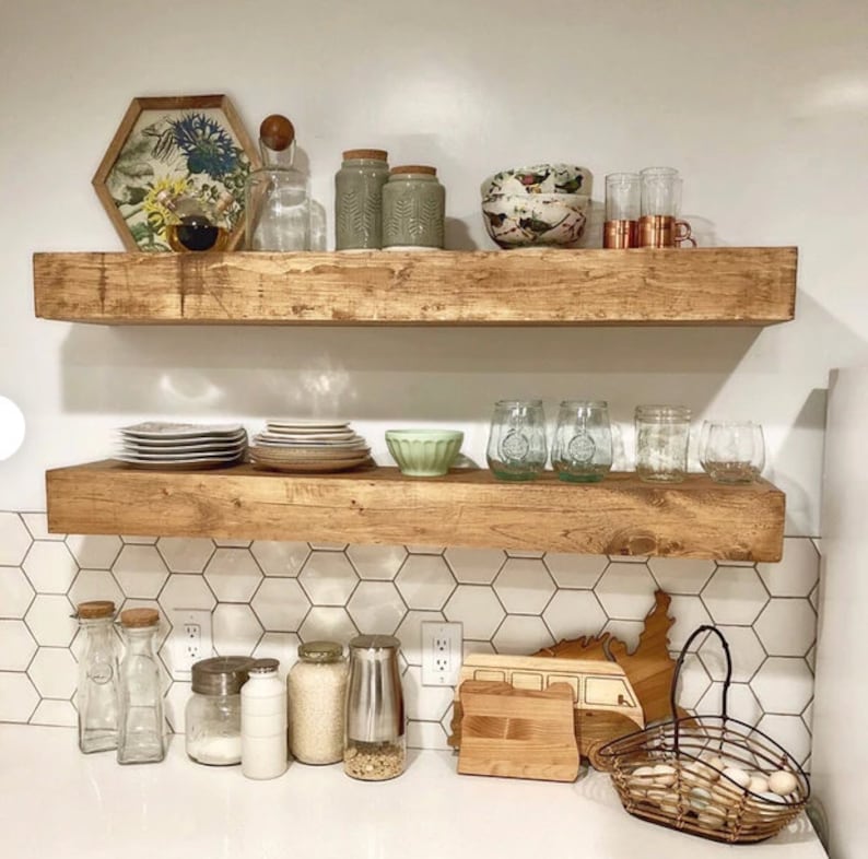 May include: Two rustic, wooden floating shelves display kitchenware and decor. Items include plates, bowls, jars, glasses, and a framed floral print. The shelves are mounted on a white wall with hexagonal tile backsplash, creating a warm, inviting kitchen space.
