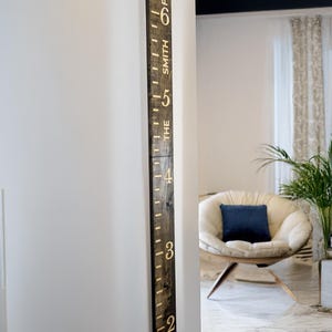 May include: A dark brown wooden height chart ruler fixed to a white wall. The ruler features gold-coloured numbers and markings, with the text "THE SMITH FAMILY" vertically aligned. The numbers range from 61 cm to 183 cm.