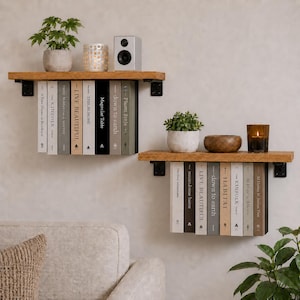 May include: Two wooden floating shelves with black metal brackets are mounted on a light-coloured wall. Each shelf holds a collection of books, small potted plants, and decorative items like candles and a speaker. The shelves create a minimalist, modern aesthetic.