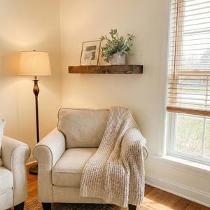 May include: A cozy living room scene featuring a beige armchair with a knitted throw blanket. A dark wooden shelf holds framed art and greenery. A floor lamp with a beige shade and a woven rug complete the warm, inviting space.