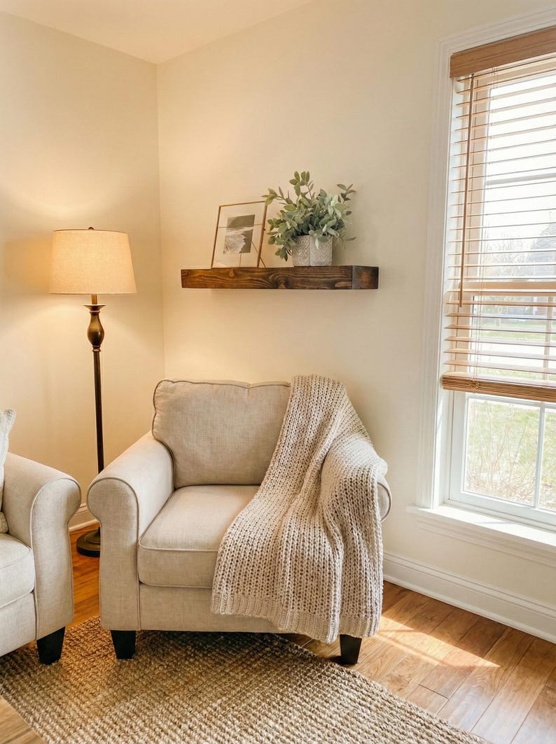 May include: A cozy living room scene featuring a beige armchair with a knitted throw blanket. A dark wooden shelf holds framed art and greenery. A floor lamp with a beige shade and a woven rug complete the warm, inviting space.