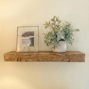 May include: A rustic, stained wooden shelf mounted on a cream-colored wall. The shelf displays a framed black and white photograph, and a potted plant with green foliage. The shelf is a warm brown color.