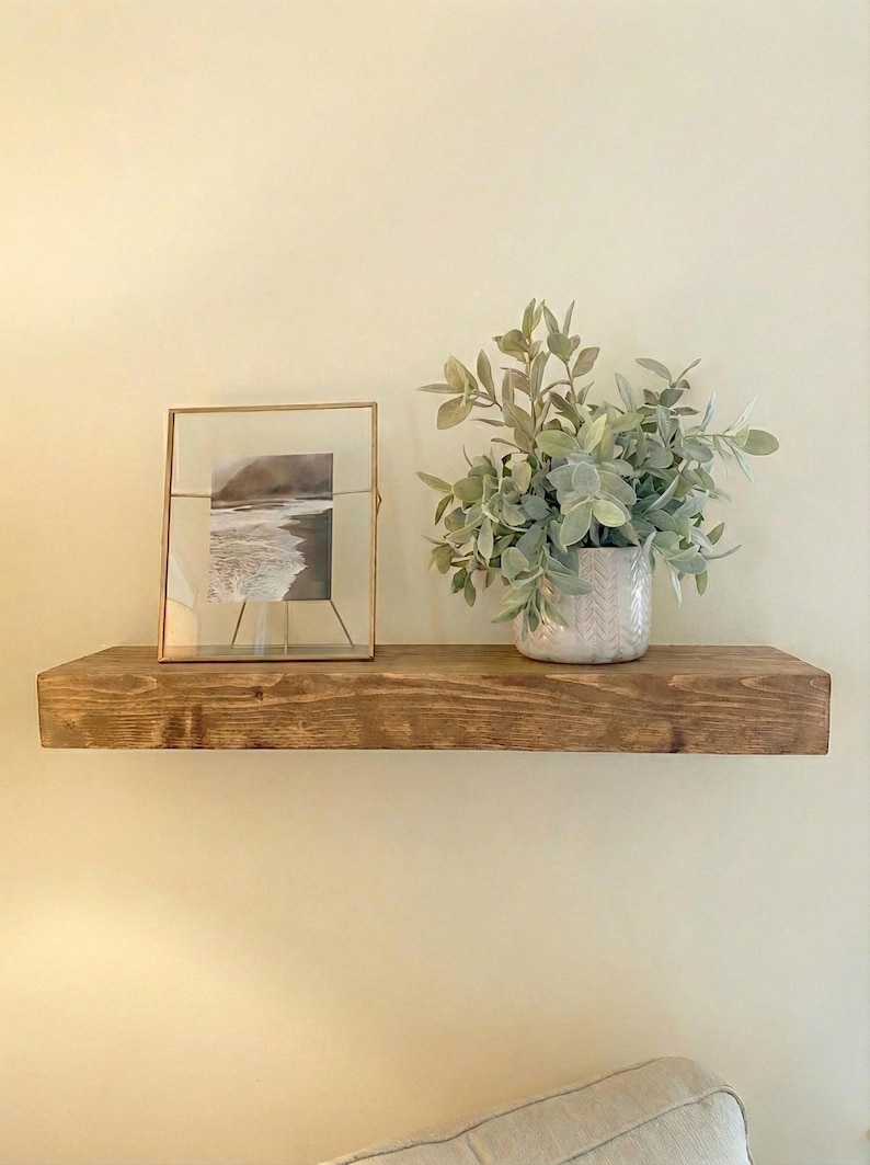 May include: A rustic, stained wooden shelf mounted on a cream-colored wall. The shelf displays a framed black and white photograph, and a potted plant with green foliage. The shelf is a warm brown color.