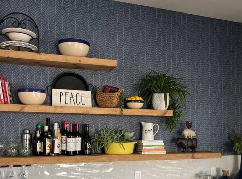 May include: Wooden shelves displaying various home decor items against a blue patterned wall. Items include bowls, plants, books, bottles of wine, and a sign that says "PEACE". The shelves are made of light-colored wood.