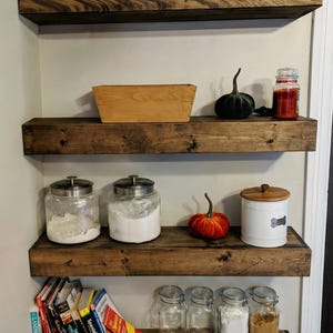 K&ouml;nnte beinhalten: Dunkel gebeizte Holz-Schweberegale mit verschiedenen K&uuml;chen- und Dekorationsartikeln. Enth&auml;lt Gl&auml;ser, K&uuml;rbisse, eine Holzkiste und B&uuml;cher. Die Regale sind braun.