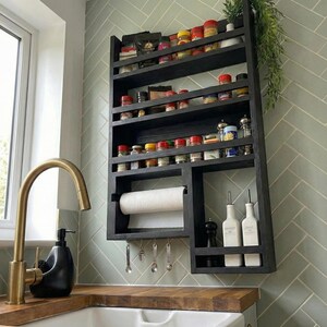 May include: A black wooden spice rack with multiple shelves, holding various spice jars. It includes a paper towel holder, small hanging spoons, and a section for oil bottles. The rack is mounted on a light green tiled wall.