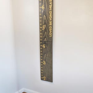 May include: A dark stained wooden height chart with gold lettering and markings. The chart is mounted on a white wall and reads "LOVED BEYOND MEASURE". The height markings are in metres, ranging from approximately 0.6 to 1.8 metres.