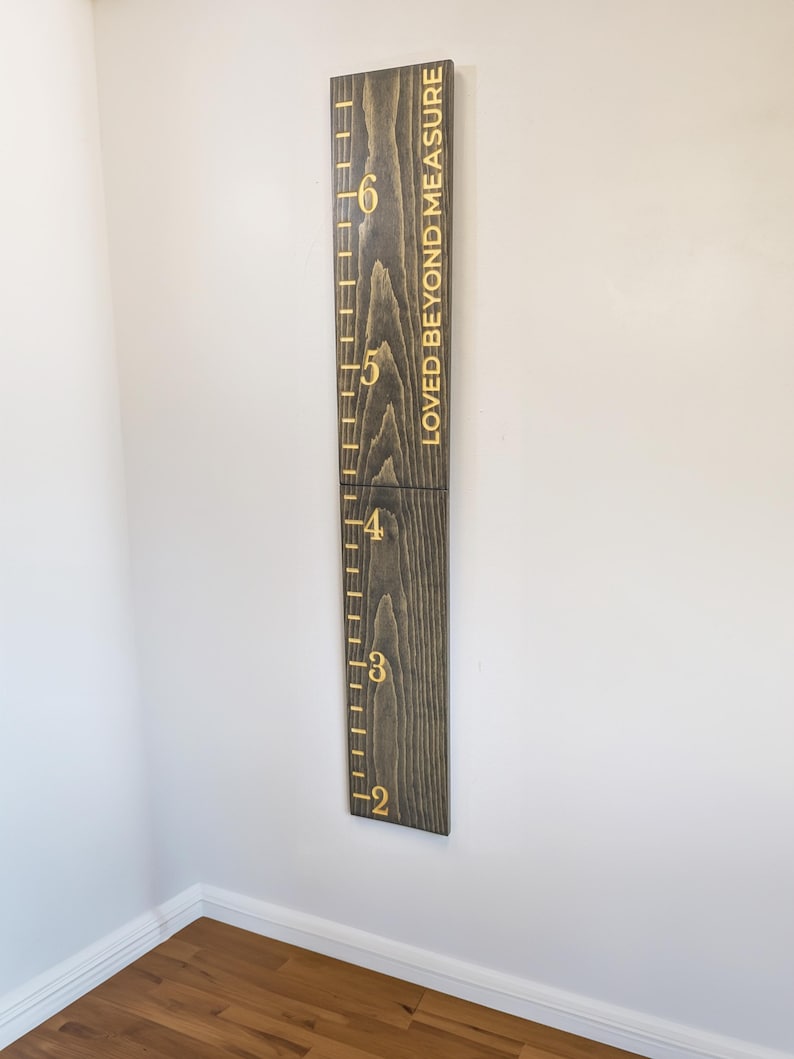 May include: A dark stained wooden height chart with gold lettering and markings. The chart is mounted on a white wall and reads "LOVED BEYOND MEASURE". The height markings are in metres, ranging from approximately 0.6 to 1.8 metres.