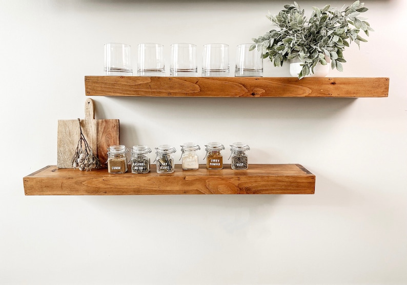 May include: Wooden floating shelves with clear drinking glasses, spice jars, a cutting board, and a potted plant. The spice jars are labeled with various herbs and spices. The shelves are stained a warm brown color.