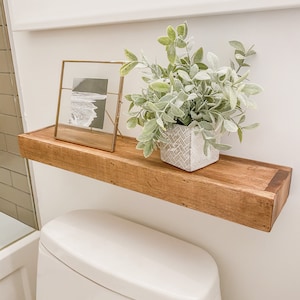 K&ouml;nnte beinhalten: Ein schwebendes Holzregal mit einem gerahmten Schwarz-Wei&szlig;-Foto und einer Topfpflanze mit gr&uuml;nen und grauen Bl&auml;ttern. Das Regal ist &uuml;ber einer wei&szlig;en Toilette in einem Badezimmer angebracht.