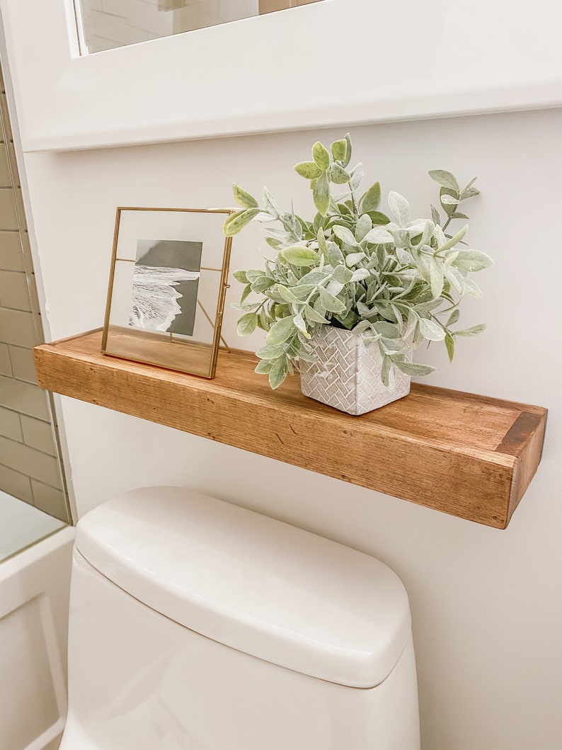 K&ouml;nnte beinhalten: Ein schwebendes Holzregal mit einem gerahmten Schwarz-Wei&szlig;-Foto und einer Topfpflanze mit gr&uuml;nen und grauen Bl&auml;ttern. Das Regal ist &uuml;ber einer wei&szlig;en Toilette in einem Badezimmer angebracht.