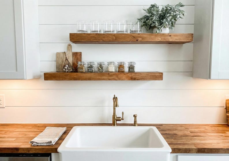 May include: A kitchen scene with wooden shelves holding clear glasses, spice jars, and decorative items. A white sink and gold faucet are in the foreground. The countertop is made of wood. The background is white.