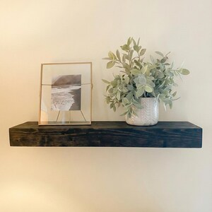 May include: A dark stained wooden floating shelf with a framed black and white photograph and a decorative vase with green foliage. The shelf is mounted on a cream-colored wall, creating a minimalist aesthetic.