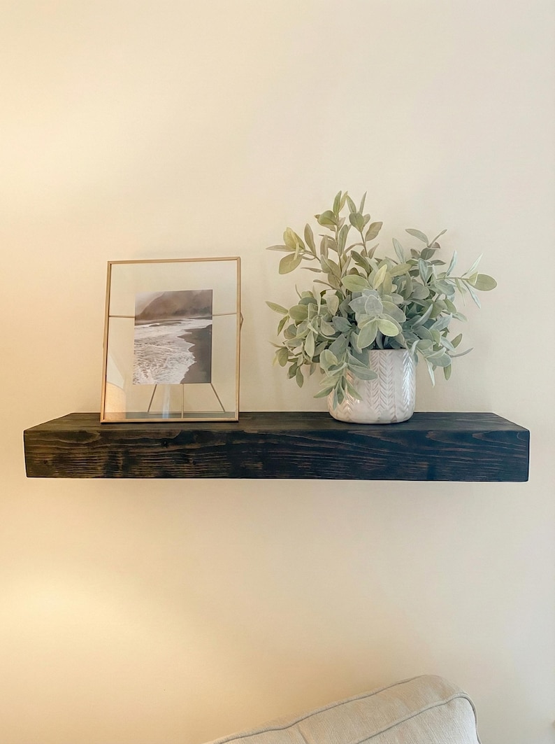 May include: A dark stained wooden floating shelf with a framed black and white photograph and a decorative vase with green foliage. The shelf is mounted on a cream-colored wall, creating a minimalist aesthetic.