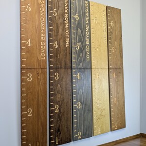 May include: A set of wooden height charts in shades of brown, featuring engraved markings and numbers. The text "LOVED BEYOND MEASURE" and family names are also engraved. These decorative charts are designed to track a child's height.