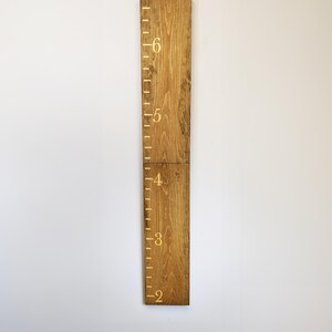 May include: A wooden height chart with gold markings and numbers from 2 to 6 feet (approximately 61 to 183 cm). The chart is made of two wooden planks joined together and mounted on a white wall. The wood has a natural grain and a warm, brown tone.