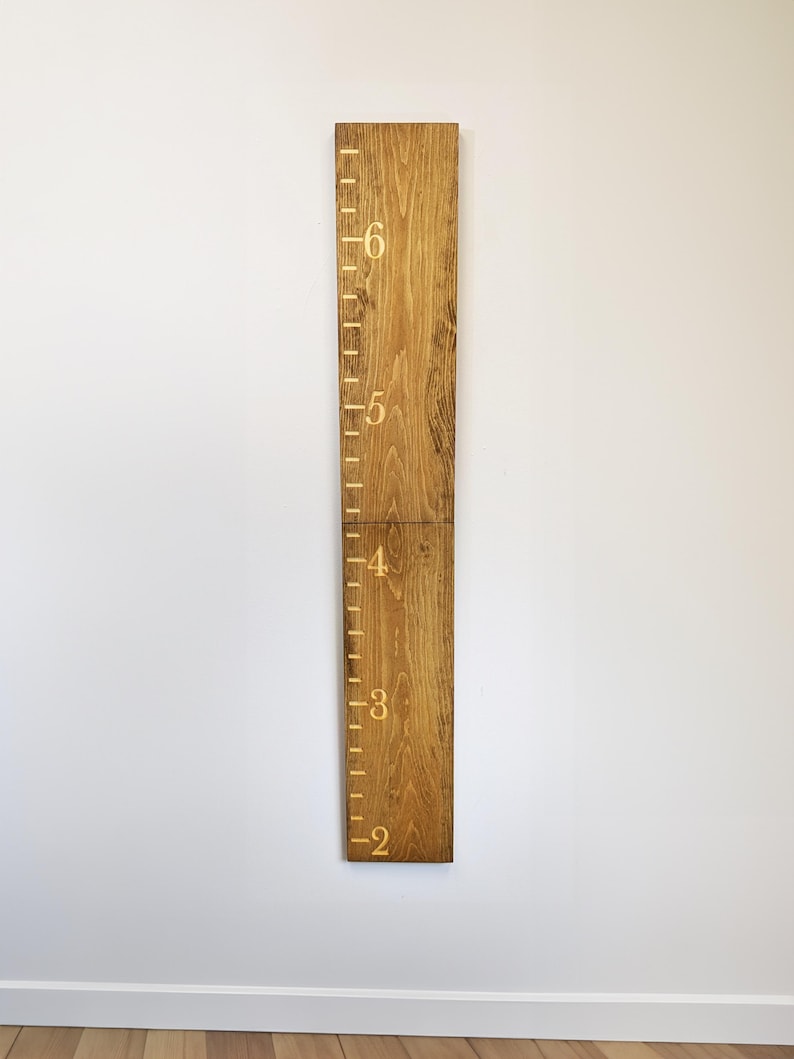 May include: A wooden height chart with gold markings and numbers from 2 to 6 feet (approximately 61 to 183 cm). The chart is made of two wooden planks joined together and mounted on a white wall. The wood has a natural grain and a warm, brown tone.