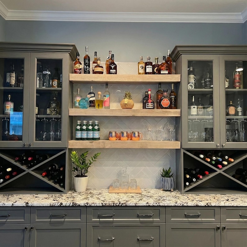 May include: A home bar setup with gray cabinets, granite countertops, and wooden shelves. The shelves display various liquor bottles, glassware, and a decorative pineapple. Wine racks are built into the cabinets, and a small plant adds a touch of greenery.