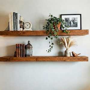 May include: Two rustic, wooden floating shelves mounted on a white wall. The top shelf holds books, a clock, and a potted plant. The bottom shelf displays books, a lantern, a vase, and a wooden bird. The shelves are stained a warm brown.
