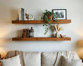 May include: Two rustic, wooden floating shelves mounted on a white wall. The top shelf holds books, a clock, and a potted plant. The bottom shelf displays books, a lantern, a vase with dried flowers, and a wooden bird figurine. A sofa is below.