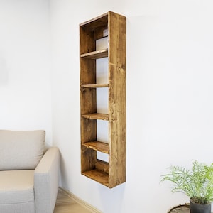 May include: A tall, narrow, wooden bookshelf with five shelves, mounted on a white wall. The bookshelf is made of stained wood, showcasing the natural grain. The shelves are evenly spaced, offering storage or display space. A sofa and a plant are in the background.