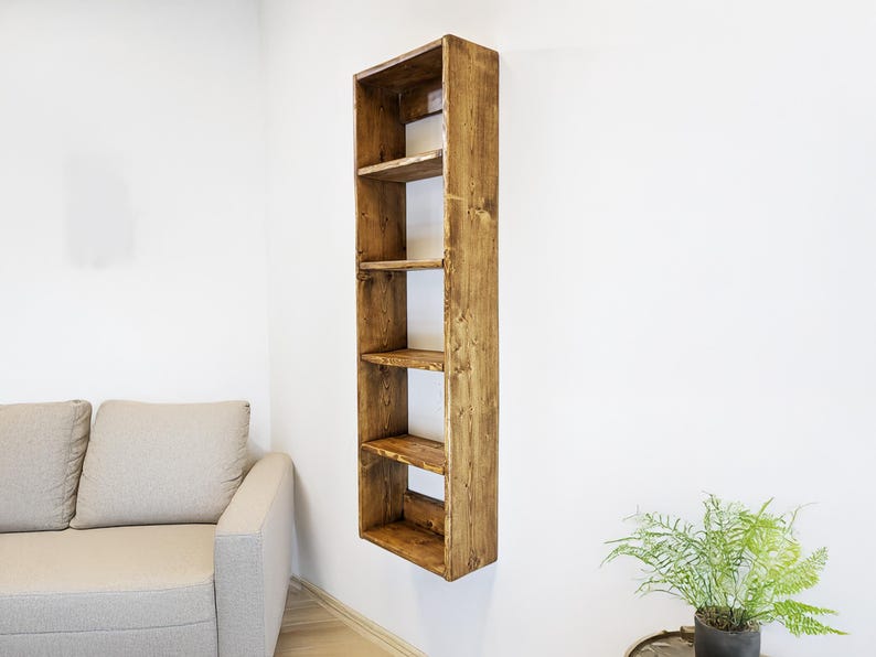 May include: A tall, narrow, wooden bookshelf with five shelves, mounted on a white wall. The bookshelf is made of stained wood, showcasing the natural grain. The shelves are evenly spaced, offering storage or display space. A sofa and a plant are in the background.