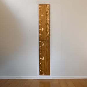 May include: A wooden height chart with the text "LOVED BEYOND MEASURE" and measurements from 1 to 5 feet (approximately 30 to 152 cm). The chart is made of light brown wood and is mounted on a white wall. The numbers and markings are in a lighter color.