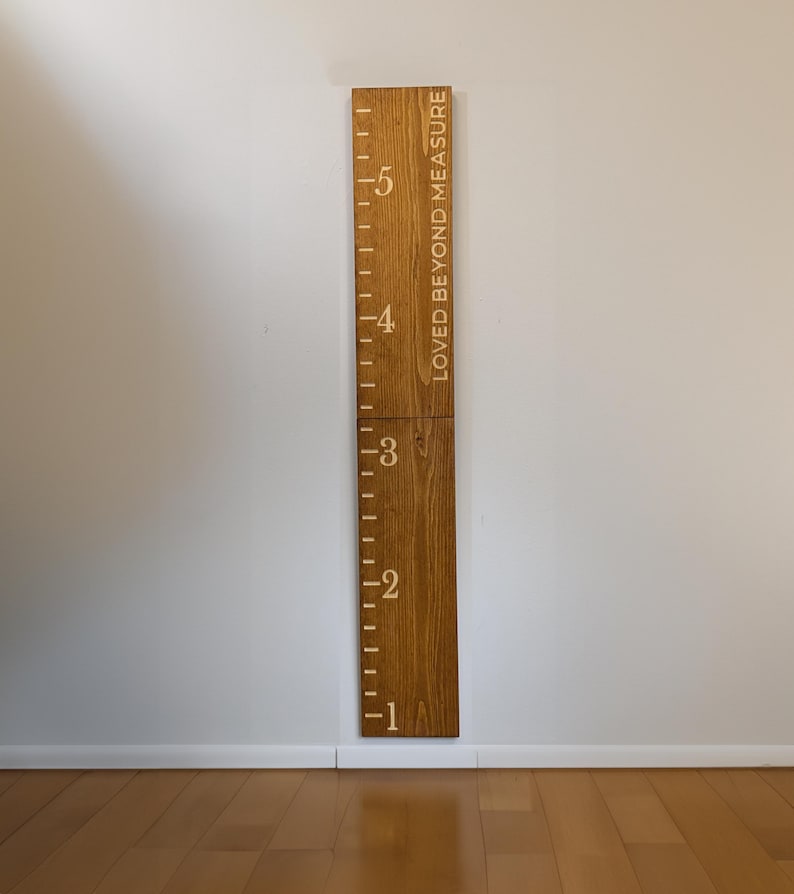 May include: A wooden height chart with the text "LOVED BEYOND MEASURE" and measurements from 1 to 5 feet (approximately 30 to 152 cm). The chart is made of light brown wood and is mounted on a white wall. The numbers and markings are in a lighter color.