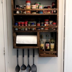 May include: A dark stained wooden spice rack with multiple shelves, holding various spice jars, cans, and bottles. A paper towel roll is mounted below the shelves, with four hanging kitchen utensils. Three bottles of wine and vinegar are also visible.