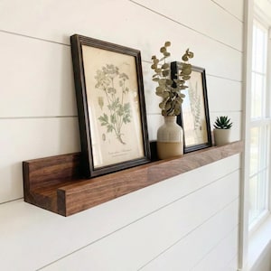 May include: A dark wood floating shelf displays framed botanical prints, a small vase with dried greenery, and a potted succulent. The shelf is mounted on a white shiplap wall, creating a minimalist, natural aesthetic.