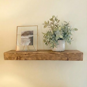May include: A rustic, stained wooden shelf mounted on a cream-colored wall. The shelf displays a framed black and white photograph, a potted plant with green foliage, and a decorative vase. The shelf is a warm brown color.