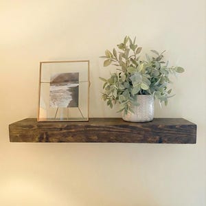 May include: A dark stained wooden floating shelf with a framed picture and a potted plant. The picture is in a gold frame and shows a black and white image of a beach. The plant is in a white patterned pot.