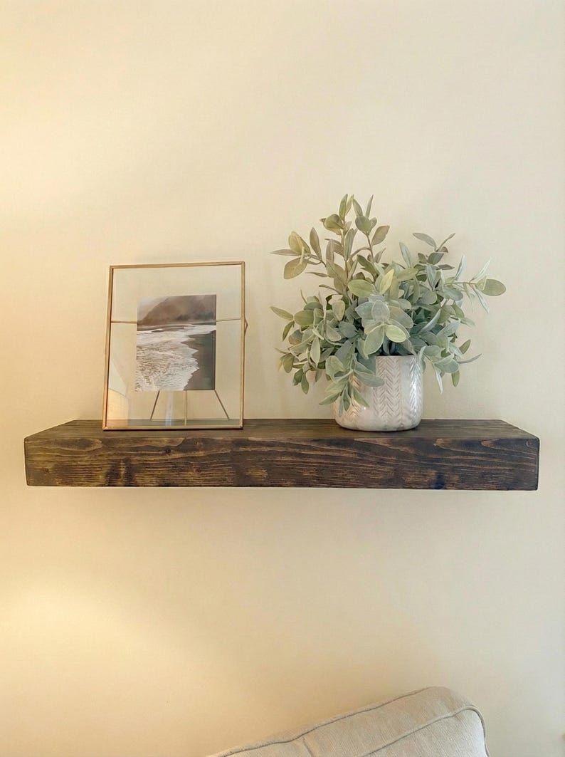 May include: A dark stained wooden floating shelf with a framed picture and a potted plant. The picture is in a gold frame and shows a black and white image of a beach. The plant is in a white patterned pot.