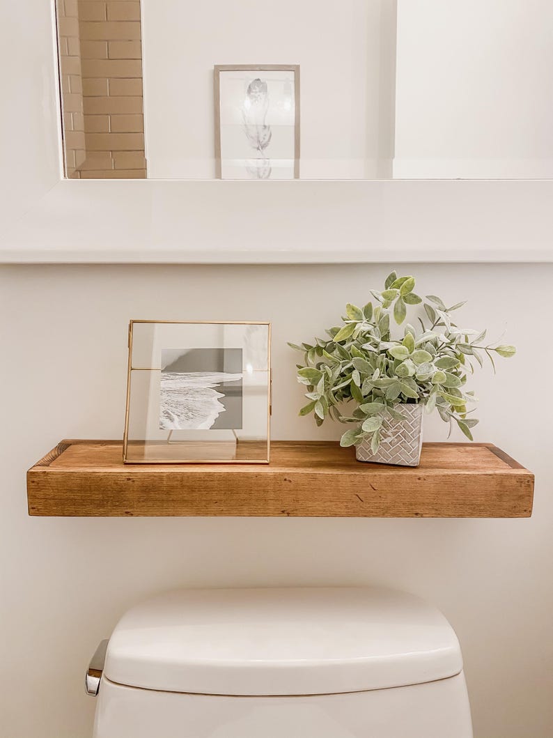 May include: A wooden floating shelf with a framed black and white photograph and a small potted plant. The shelf is mounted above a white toilet. A framed feather art piece is visible in the background.