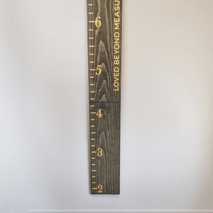 May include: A dark stained wooden height chart with gold lettering. The chart measures from 61 cm to 183 cm, with the words "LOVED BEYOND MEASURE" printed vertically. It is mounted on a white wall.