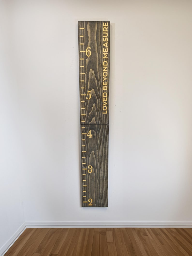 May include: A dark stained wooden height chart with gold lettering. The chart measures from 61 cm to 183 cm, with the words "LOVED BEYOND MEASURE" printed vertically. It is mounted on a white wall.