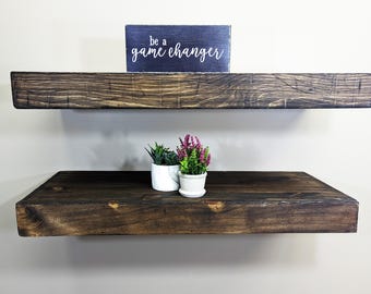 May include: Two dark stained wooden floating shelves on a light wall. The top shelf has a sign reading "be a game changer." The bottom shelf holds two small potted plants. The shelves are likely made of solid wood.