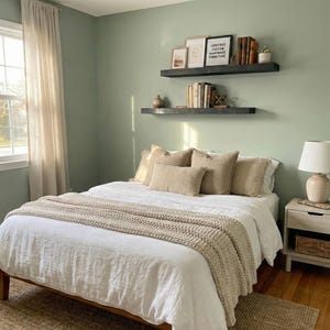 May include: A bedroom with a bed covered in white linens, beige pillows, and a chunky knit throw. Two floating shelves on the wall display books, framed art, and decorative items. A bedside table with a lamp and a woven basket is on the right.