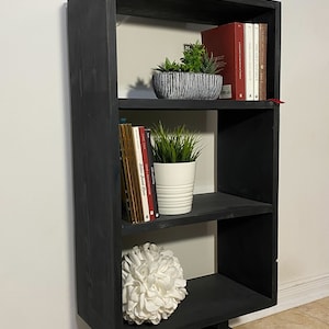 May include: A black wooden bookshelf with three shelves. The top shelf holds a framed print with text, a gray decorative flower, and a white ceramic cat with a gold crown. The shelves display books and plants.