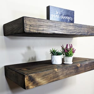 May include: Two dark stained wooden floating shelves mounted on a white wall. The top shelf has a small black sign that reads "be a game changer." The bottom shelf holds two small potted plants.