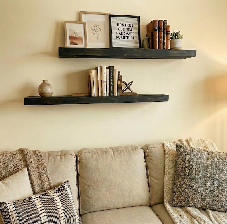 May include: Two dark stained wooden floating shelves mounted on a cream-colored wall. The top shelf holds framed art, books, and a small plant. The bottom shelf displays books and a decorative vase. The text on the top shelf reads "IRNTREE CUSTOM HANDMADE FURNITURE."