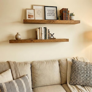 May include: Two rustic, dark-stained wooden floating shelves mounted on a cream-colored wall. The top shelf holds framed artwork, books, and a small plant. The bottom shelf displays books and a decorative vase. The text on the sign reads: IIRNTREE CUSTOM HANDMADE FURNITURE.