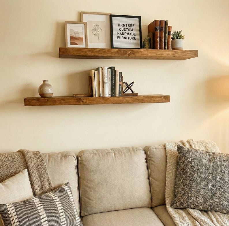 May include: Two rustic, dark-stained wooden floating shelves mounted on a cream-colored wall. The top shelf holds framed artwork, books, and a small plant. The bottom shelf displays books and a decorative vase. The text on the sign reads: IIRNTREE CUSTOM HANDMADE FURNITURE.