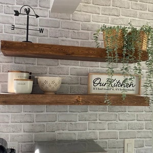 May include: Two rustic, stained wooden shelves mounted on a brick-patterned wall. The top shelf holds two woven baskets with trailing greenery. The bottom shelf displays a sign that reads "Our Kitchen" and two ceramic containers.